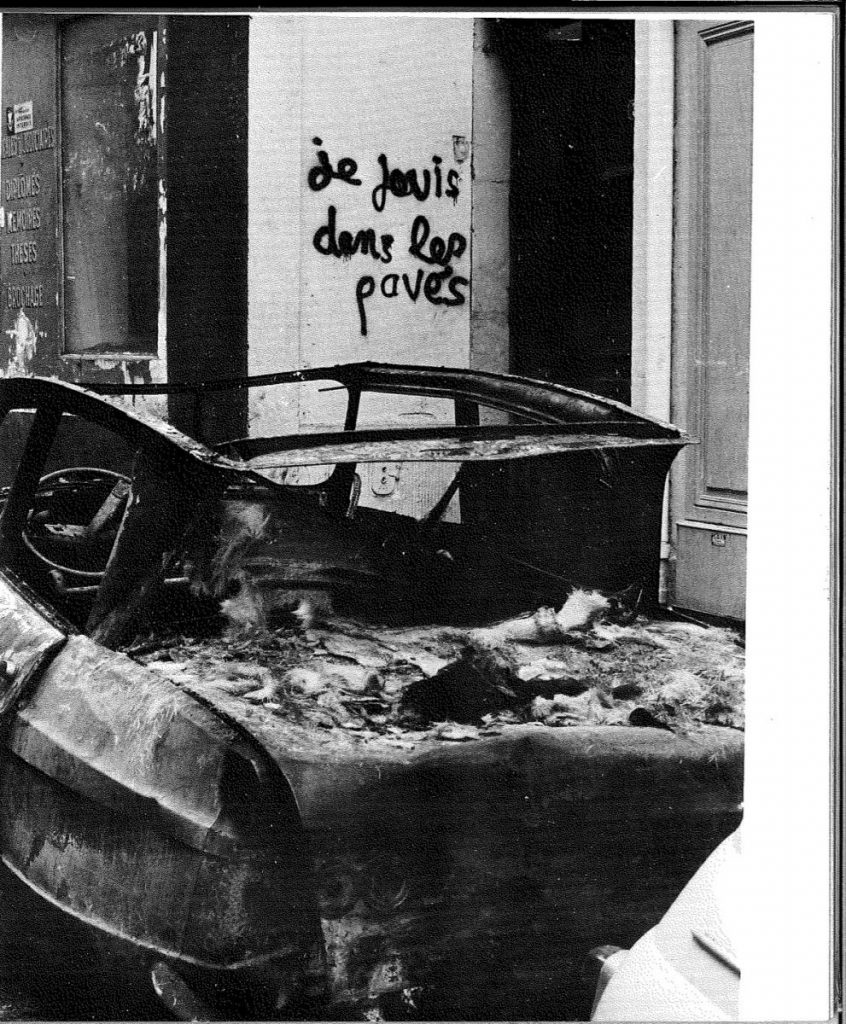 Black and white photo shows the wreckage of a car in the foreground and graffiti in the background reading 'je sevis dans les pavés'