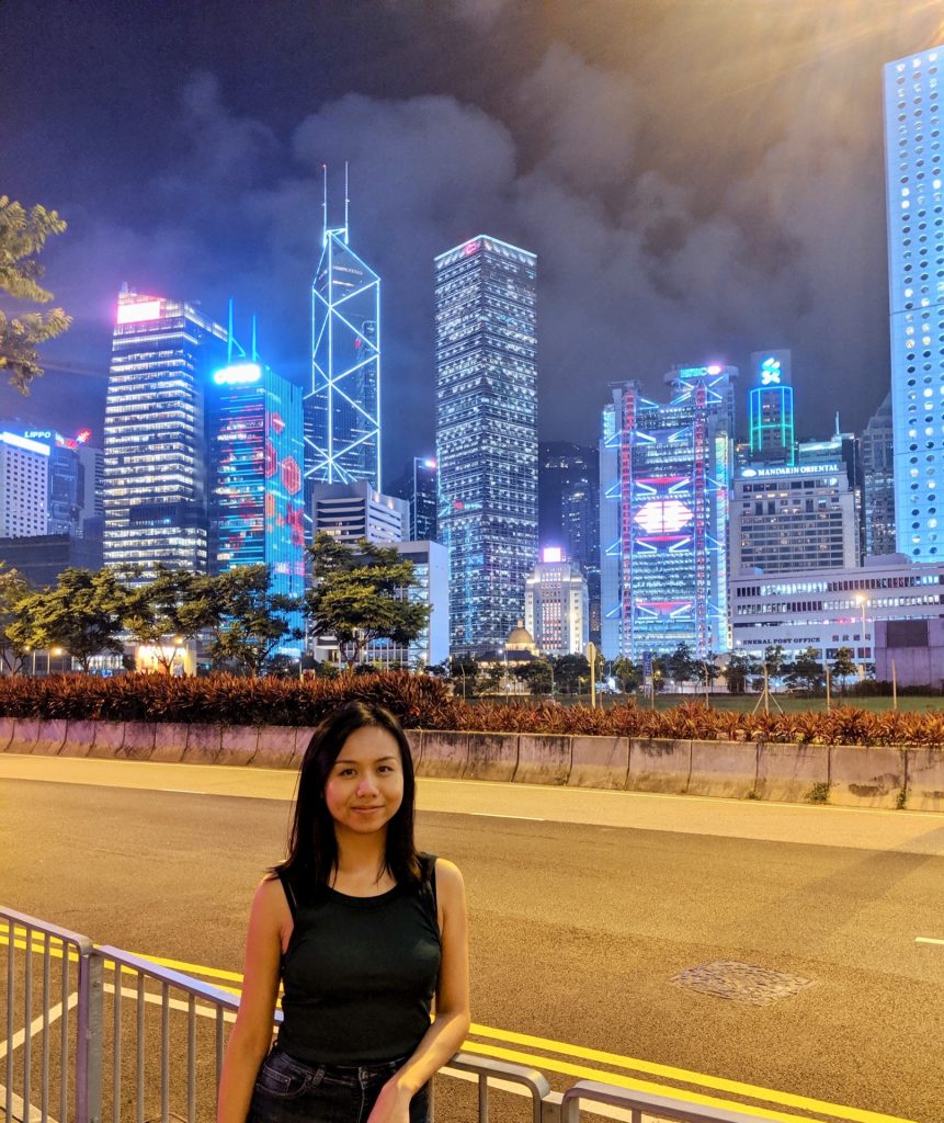 Image shows a cityscape at night in the background, lit up skyscrapers. Vivian stands in the foreground.