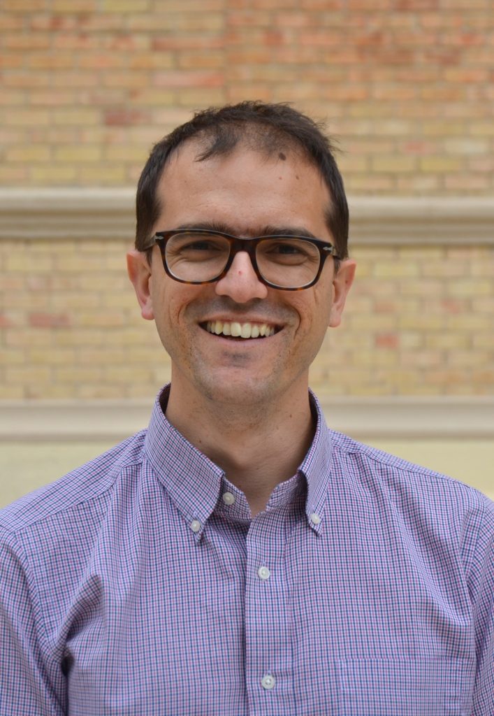 Portrait picture of Lorenzo Costaguta, smiling, wearing glasses and a shirt in front of a brick wall